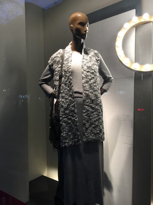 A mannequin is dressed in a long gray dress with a textured, sleeveless cardigan. It stands in a shop window with a circular light fixture on the wall nearby, casting a warm glow. A dark-brown handbag is slung over the mannequin's left shoulder.