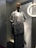 A mannequin is dressed in a long gray dress with a textured, sleeveless cardigan. It stands in a shop window with a circular light fixture on the wall nearby, casting a warm glow. A dark-brown handbag is slung over the mannequin's left shoulder.