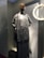 A mannequin is dressed in a long gray dress with a textured, sleeveless cardigan. It stands in a shop window with a circular light fixture on the wall nearby, casting a warm glow. A dark-brown handbag is slung over the mannequin's left shoulder.