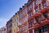 A row of colorful, ornate European-style apartment buildings with intricate architectural details such as balconies and decorative elements. The buildings are painted in shades of red, yellow, and pink, standing against a clear blue sky.