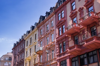 A row of colorful, ornate European-style apartment buildings with intricate architectural details such as balconies and decorative elements. The buildings are painted in shades of red, yellow, and pink, standing against a clear blue sky.