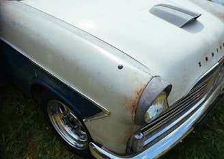 A vintage car features a hood with a scoop and visible rust spots on the off-white body. The word 'Zodiac' is visible on the front near the headlight, which is surrounded by chrome detailing. The car is parked on grass, and the wheel hubcaps are shiny.
