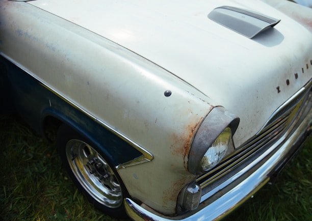 A vintage car features a hood with a scoop and visible rust spots on the off-white body. The word 'Zodiac' is visible on the front near the headlight, which is surrounded by chrome detailing. The car is parked on grass, and the wheel hubcaps are shiny.