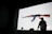 A large image of a rifle is projected onto a screen with a person in a dark hoodie standing in front of it, casting a shadow. A camera is set on a tripod next to an empty chair in a dimly lit room.
