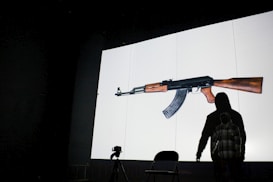 A large image of a rifle is projected onto a screen with a person in a dark hoodie standing in front of it, casting a shadow. A camera is set on a tripod next to an empty chair in a dimly lit room.