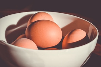 A bunch of brown eggs rest gently in a white ceramic bowl. The lighting creates soft shadows and highlights, giving the eggs a smooth, matte appearance. The background is dark, drawing focus to the bowl of eggs.