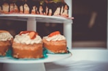 A close-up view of delicious desserts displayed on a two-tiered platter. The top row features a cake with a chocolate drizzle, while the bottom row showcases individual mini cakes topped with whipped cream and fresh fruit slices. The background is softly blurred, bringing focus to the desserts.