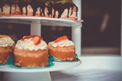 A close-up view of delicious desserts displayed on a two-tiered platter. The top row features a cake with a chocolate drizzle, while the bottom row showcases individual mini cakes topped with whipped cream and fresh fruit slices. The background is softly blurred, bringing focus to the desserts.