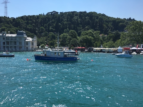 A blue boat is floating on a vibrant turquoise body of water, surrounded by several small boats. In the background, there is a lush green hillside and a grand white building. The shoreline is dotted with trees and tents, suggesting a lively waterfront area.