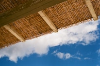 A bamboo roof or canopy stretches across the top of the frame against a backdrop of a clear blue sky with scattered white clouds.