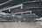 A spacious and modern airport terminal features a high, curved ceiling with visible steel beams and large air ducts. The terminal floor is wide and open, with groups of people visible in the distance near check-in counters and signage. Large windows allow natural light to illuminate the space.