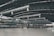 A spacious and modern airport terminal features a high, curved ceiling with visible steel beams and large air ducts. The terminal floor is wide and open, with groups of people visible in the distance near check-in counters and signage. Large windows allow natural light to illuminate the space.