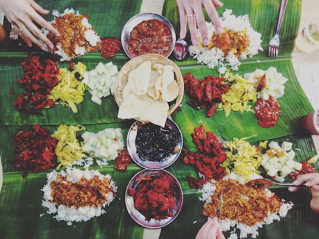 A communal meal is spread out on banana leaves, featuring a variety of colorful dishes including rice, curries, vegetables, and relishes. Several hands are reaching in to partake in the meal, suggesting a shared dining experience. The central basket contains crispy, round snacks.