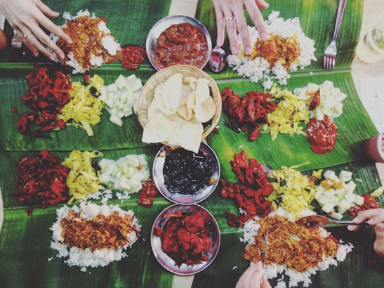 A communal meal is spread out on banana leaves, featuring a variety of colorful dishes including rice, curries, vegetables, and relishes. Several hands are reaching in to partake in the meal, suggesting a shared dining experience. The central basket contains crispy, round snacks.
