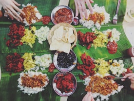 A communal meal is spread out on banana leaves, featuring a variety of colorful dishes including rice, curries, vegetables, and relishes. Several hands are reaching in to partake in the meal, suggesting a shared dining experience. The central basket contains crispy, round snacks.