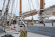 Numerous ropes and rigging are prominently displayed across the deck of an old sailboat. The wooden masts and rolled up sails dominate the scene, with equipment like winches and ropes organized and secured. A lifebuoy, wooden benches, and a fire extinguisher can also be seen on the deck, suggesting readiness for maritime travel.