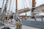 Numerous ropes and rigging are prominently displayed across the deck of an old sailboat. The wooden masts and rolled up sails dominate the scene, with equipment like winches and ropes organized and secured. A lifebuoy, wooden benches, and a fire extinguisher can also be seen on the deck, suggesting readiness for maritime travel.