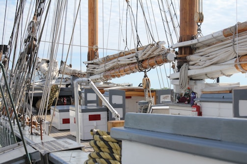 Numerous ropes and rigging are prominently displayed across the deck of an old sailboat. The wooden masts and rolled up sails dominate the scene, with equipment like winches and ropes organized and secured. A lifebuoy, wooden benches, and a fire extinguisher can also be seen on the deck, suggesting readiness for maritime travel.