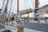 Numerous ropes and rigging are prominently displayed across the deck of an old sailboat. The wooden masts and rolled up sails dominate the scene, with equipment like winches and ropes organized and secured. A lifebuoy, wooden benches, and a fire extinguisher can also be seen on the deck, suggesting readiness for maritime travel.