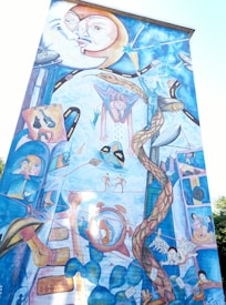 A vibrant mural featuring surreal imagery including a large moon with faces, various abstract human figures, musical instruments, and objects like a telephone, light bulb, and flying wings. The composition appears whimsical with a blend of different elements, such as swirling patterns and a twisting vine that runs vertically through the artwork.