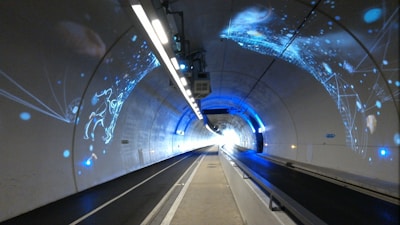 A tunnel with smooth, curved walls features dynamic blue and white digital projections, creating a futuristic atmosphere. The projections form abstract, geometric shapes and constellations against the tunnel's clean surfaces, while bright linear lights run along the ceiling.