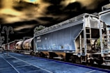 A train with several freight cars moves along railway tracks. The visible cars include a large, gray hopper car and a cylindrical tank car. The scene has a surreal, high-contrast effect with dark skies and vivid outlines. A platform is visible in the foreground, accompanied by street lamps on the left side.