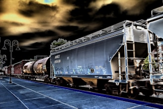 A train with several freight cars moves along railway tracks. The visible cars include a large, gray hopper car and a cylindrical tank car. The scene has a surreal, high-contrast effect with dark skies and vivid outlines. A platform is visible in the foreground, accompanied by street lamps on the left side.