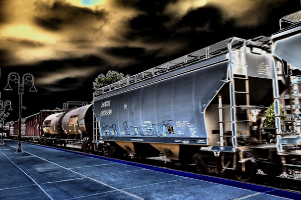 A train with several freight cars moves along railway tracks. The visible cars include a large, gray hopper car and a cylindrical tank car. The scene has a surreal, high-contrast effect with dark skies and vivid outlines. A platform is visible in the foreground, accompanied by street lamps on the left side.