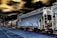 A train with several freight cars moves along railway tracks. The visible cars include a large, gray hopper car and a cylindrical tank car. The scene has a surreal, high-contrast effect with dark skies and vivid outlines. A platform is visible in the foreground, accompanied by street lamps on the left side.