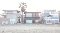 Three modern beach houses with large windows and balconies are situated side by side. A sandy beach lies in the foreground, and a tall palm tree stands behind one of the houses. Bright red umbrellas are visible on the patios, adding a pop of color to the neutral tones of the buildings.