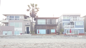 Three modern beach houses with large windows and balconies are situated side by side. A sandy beach lies in the foreground, and a tall palm tree stands behind one of the houses. Bright red umbrellas are visible on the patios, adding a pop of color to the neutral tones of the buildings.
