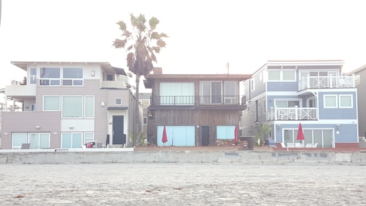 Three modern beach houses with large windows and balconies are situated side by side. A sandy beach lies in the foreground, and a tall palm tree stands behind one of the houses. Bright red umbrellas are visible on the patios, adding a pop of color to the neutral tones of the buildings.