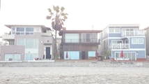 Three modern beach houses with large windows and balconies are situated side by side. A sandy beach lies in the foreground, and a tall palm tree stands behind one of the houses. Bright red umbrellas are visible on the patios, adding a pop of color to the neutral tones of the buildings.