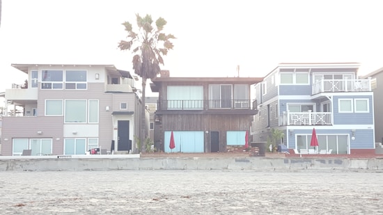 Three modern beach houses with large windows and balconies are situated side by side. A sandy beach lies in the foreground, and a tall palm tree stands behind one of the houses. Bright red umbrellas are visible on the patios, adding a pop of color to the neutral tones of the buildings.