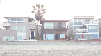 Three modern beach houses with large windows and balconies are situated side by side. A sandy beach lies in the foreground, and a tall palm tree stands behind one of the houses. Bright red umbrellas are visible on the patios, adding a pop of color to the neutral tones of the buildings.