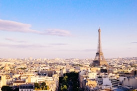 A panoramic view of Paris with the Eiffel Tower prominently visible against a clear blue sky. The cityscape is filled with classic Parisian architecture, featuring a mix of old and modern buildings. Soft clouds are scattered in the sky, adding to the serene atmosphere.