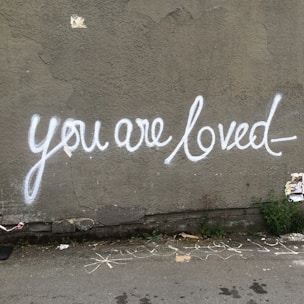 Graffiti with the message 'you are loved' is painted in white on a textured grey wall. The script is stylized and flows smoothly across the surface. Some greenery and torn paper are visible at the base of the wall, along with a few chalk markings on the ground.