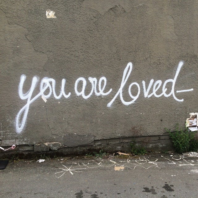 Graffiti with the message 'you are loved' is painted in white on a textured grey wall. The script is stylized and flows smoothly across the surface. Some greenery and torn paper are visible at the base of the wall, along with a few chalk markings on the ground.
