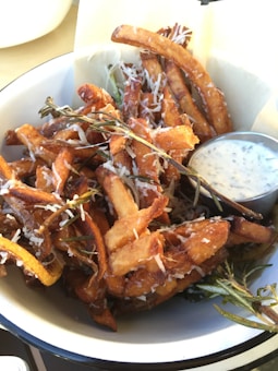 Crispy sweet potato fries topped with grated cheese and garnished with sprigs of rosemary are served in a white bowl. A small container of creamy dipping sauce accompanies the fries.