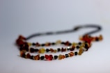 A necklace composed of small, irregularly shaped beads in shades of amber, red, and black rests on a plain surface. The beads are strung together, forming a natural and earthy aesthetic with variations in color and texture.