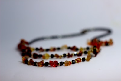 A necklace composed of small, irregularly shaped beads in shades of amber, red, and black rests on a plain surface. The beads are strung together, forming a natural and earthy aesthetic with variations in color and texture.
