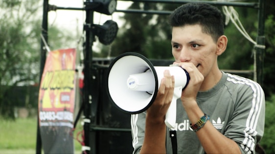 A person is holding a white megaphone close to their face, appearing to speak into it. They are wearing a gray Adidas sweatshirt with white stripes on the sleeves. In the background, there is some audio or stage equipment, along with a colorful poster. The setting seems to be outdoors.