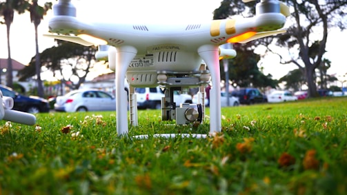 A drone positioned low to the ground on vibrant green grass. The focus is on the drone's camera and undercarriage, with blurred trees and parked cars in the background, suggesting the setting is a park or public area.