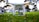 A drone positioned low to the ground on vibrant green grass. The focus is on the drone's camera and undercarriage, with blurred trees and parked cars in the background, suggesting the setting is a park or public area.