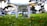 A drone positioned low to the ground on vibrant green grass. The focus is on the drone's camera and undercarriage, with blurred trees and parked cars in the background, suggesting the setting is a park or public area.