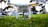 A drone positioned low to the ground on vibrant green grass. The focus is on the drone's camera and undercarriage, with blurred trees and parked cars in the background, suggesting the setting is a park or public area.