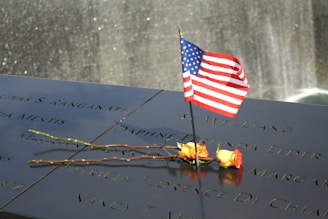 A smooth, polished memorial surface features engraved names with a small American flag and two yellow roses placed on it. In the background, a waterfall shimmers subtly.