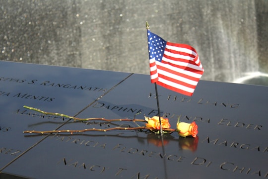 A smooth, polished memorial surface features engraved names with a small American flag and two yellow roses placed on it. In the background, a waterfall shimmers subtly.