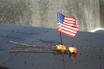 A smooth, polished memorial surface features engraved names with a small American flag and two yellow roses placed on it. In the background, a waterfall shimmers subtly.