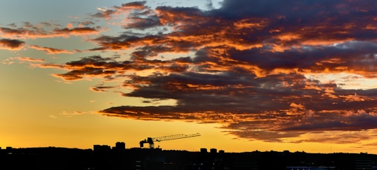 A dramatic sunset with vibrant orange and red hues illuminating the clouds above a city skyline. A construction crane is silhouetted against the colorful sky, enhancing the urban feel.